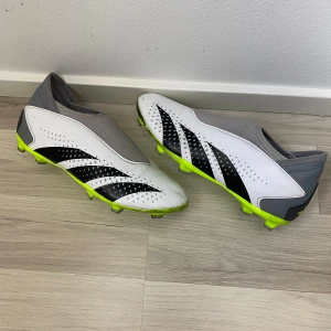 Adidas Predator fotbollsskor - Snygga Adidas Predator fotbollsskor i vitt och grått med svarta och neongula detaljer. Skorna har en strömlinjeformad design utan snörning och är perfekta för snabba rörelser på planen. Tillverkade i syntetmaterial för hållbarhet och komfort. Använda på konstgräsplan och därför det är svarta remor, inga skador eller defekter. Användes ungefär 2 veckor tills jag slutade i fotboll.