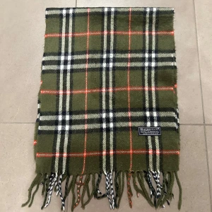 Grön rutig halsduk från Burberry - Säljer en klassisk grön rutig halsduk från Burberry, tillverkad i 100% lammull. Den har fransar i ändarna och ett tidlöst mönster i svart, vitt och rött. Perfekt för att hålla dig varm och stilren under kyliga dagar.