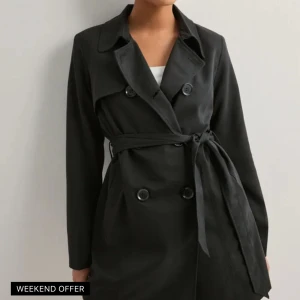 Svart trenchcoat från ONLY - Snygg svart trenchcoat från ONLY med knappar och knytskärp i midjan. Jackan har en klassisk krage och långa ärmar, perfekt för en stilren look. Passar utmärkt till både vardag och mer uppklädda tillfällen.