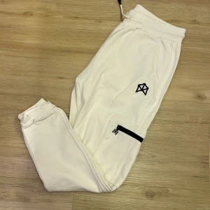 Vita mjukisbyxor från Rawgear - Snygga vita mjukisbyxor från Rawgear med svarta dragkedjor och broderad logga. Byxorna har elastisk midja med snörning och muddar vid bensluten. Perfekta för en avslappnad stil.