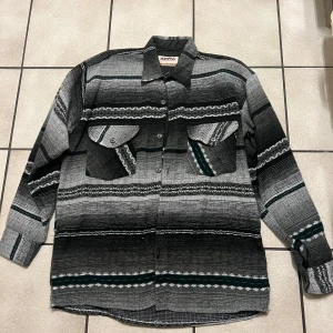 Randig skjorta från Arma Collection - Säljer en cool randig skjorta från Arma Collection i svart, grått och vitt. Skjortan har långa ärmar och två bröstfickor med knappar. Perfekt för en avslappnad stil.