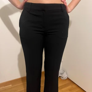 Svarta kostymbyxor - Snygga svarta kostymbyxor med medelhög midja. Perfekta för en stilren look. Byxorna har diskreta fickor och en smidig dragkedja framtill.