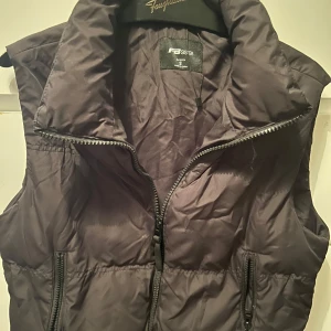 Svart dunväst - Skriv för bild på. Svart dunväst från FB Sister i storlek S. Västen har en dragkedja framtill och två praktiska fickor med dragkedja.