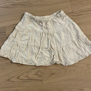 Beige volangkjol - Säljer en söt beige kortkjol med volanger. Kjolen har en elastisk midja och är perfekt för varma dagar. Den är gjord i ett lätt och luftigt material som ger en avslappnad look. Storlek: XS