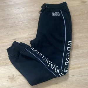 Svarta mjukisbyxor från Black Squad - Snygga svarta mjukisbyxor från Black Squad med texten 'WORLDWIDE' längs benen. Byxorna har en elastisk midja med dragsko och muddar vid bensluten. Perfekta för en avslappnad stil.