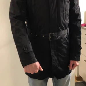 Svart trenchcoat från J.Lindeberg - Säljer en stilren svart trenchcoat från J.Lindeberg. Jackan har ett bälte i midjan och långa ärmar. Perfekt för höst och vår med sin blandning av bomull och nylon. Den har knappar och en diskret design som passar till många tillfällen.
