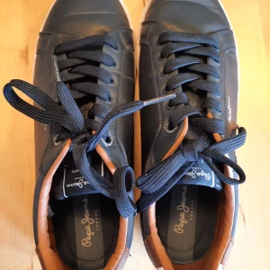 Mörkblå sneakers från Pepe Jeans - Snygga mörkblå sneakers från Pepe Jeans med bruna detaljer och vit sula. Skorna har snörning och en stilren design med märkets logga på sidan och hälen. Perfekta för en avslappnad stil.