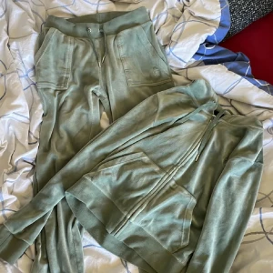 Gröna mjukisbyxor och kofta från Juicy Couture - Snygga gröna mjukisbyxor och kofta från Juicy Couture med dragkedja och fickor med knappar. Perfekta för en avslappnad stil. Byxorna och koftan har en bekväm passform och är tillverkade i ett mjukt material. 