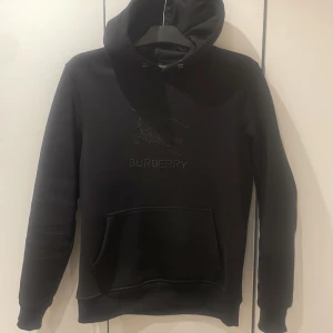 Svart hoodie från Burberry - Säljer en stilren svart hoodie från Burberry med broderad logga på bröstet. Tröjan har en klassisk känguruficka och en bekväm huva. Perfekt för en avslappnad och trendig look.