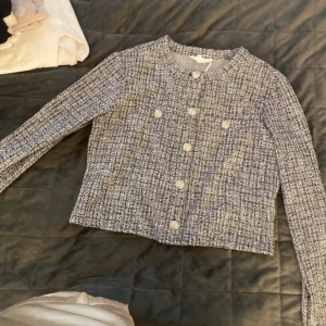 Grå tweedjacka med knappar - Snygg grå tweedjacka med rund hals och knappar framtill. Jackan har en klassisk look med långärmade ärmar och en rak passform. Perfekt för en stilren outfit.