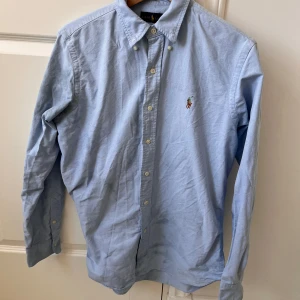 Blå skjorta från Ralph Lauren - Säljer en klassisk ljusblå skjorta från Ralph Lauren med broderad logotyp på bröstet. Skjortan har långa ärmar och knappar framtill. Perfekt för en stilren look. Väldigt missfärgad under vänstra armen. 