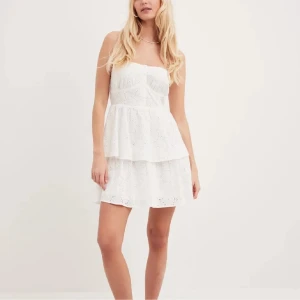 Vit axelbandslös klänning - vit axelbandslös klänning från nakd,med broderade detaljer och volang. Klänningen har en kort längd och en söt, luftig passform. Endast använd 1 gång.