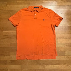 Orange pikétröja från Ralph Lauren - Säljer en snygg orange pikétröja från Ralph Lauren med klassisk krage och knappar. Tröjan har en broderad blå logga på bröstet. Perfekt för en stilren look! 