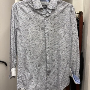 Vit blommig skjorta - Säljer en stilren vit skjorta med ett diskret blommigt mönster i blått. Skjortan har långa ärmar och klassisk krage, perfekt för en uppklädd look. Passar bra till både jeans och kostymbyxor. Perfekt för vår och sommar!