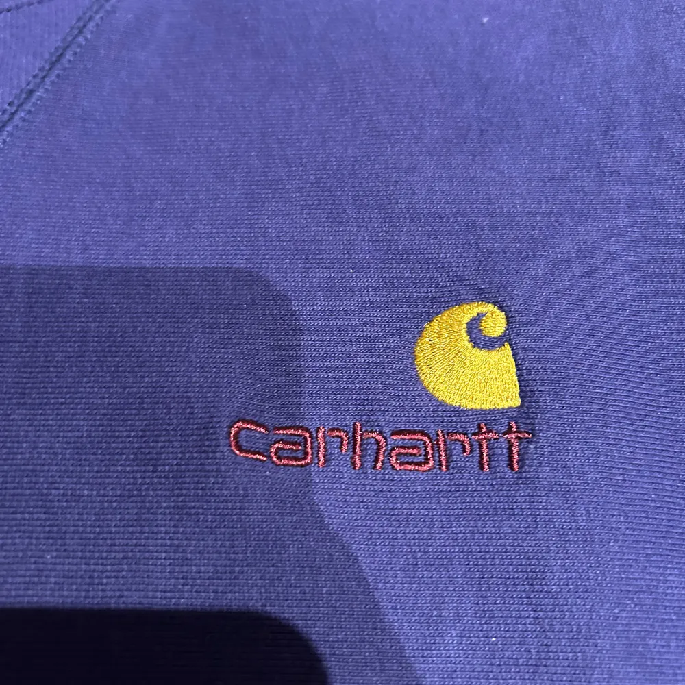 Säljer en snygg mörkblå sweatshirt från Carhartt med broderad logga i gult och rött på bröstet. Tröjan har en klassisk rund hals och långa ärmar. Perfekt för en avslappnad stil.. Neuletakit & Villapaidat.