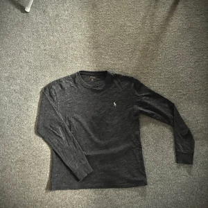 Mörkgrå långärmad tröja från Ralph Lauren - Säljer en stilren mörkgrå långärmad tröja från Ralph Lauren med det klassiska logotypbroderiet på bröstet. Perfekt för en avslappnad och snygg look. Tröjan har en rund halsringning och är tillverkad i ett mjukt material. När bilden tas är tröjan lite blöt på magen syns om man kollar noga på bilden.