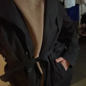Svart trenchcoat med bälte - Svart trenchcoat med knytbälte i midjan. Jackan har långa ärmar och en klassisk krage. Vårjacka. 