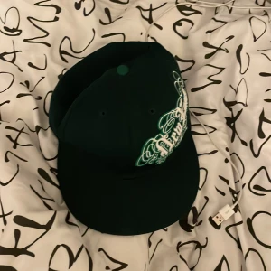 Grön ny keps - Snygg grön keps från New Era med broderad text 'New York' i vitt på framsidan. Kepsen har en platt skärm och justerbar passform. Perfekt för att ge din outfit en cool touch. Priset kan sänkas vid snabb affär