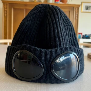 Svart ribbad mössa från C.P. Company - Snygg svart ribbad mössa från C.P. Company med unika glasögondetaljer framtill. Perfekt för att hålla värmen och samtidigt se stilren ut. En storlek som passar de flesta. Använd endast ett par tillfällen. 