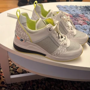 Vita sneakers med gul detalj - Snygga vita sneakers från Michael Kors med gul insida och detaljer. Skorna har ett diskret mönster och snörning framtill. Perfekta för en stilren look med en touch av färg.