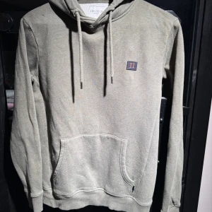 Grön hoodie från Les Deux - Säljer en stilren grön hoodie från Les Deux med en liten logga på bröstet. Tröjan har en klassisk känguruficka och justerbar huva med dragsko. Perfekt för en avslappnad stil.