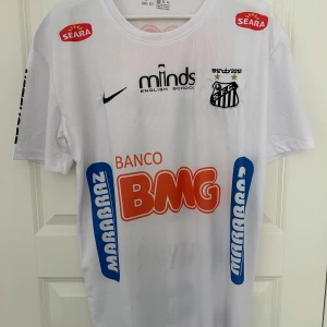 Vit fotbollströja från Nike - Säljer en vit fotbollströja från Nike med Santos FC-logga och flera sponsormärken som BMG och Seara. Tröjan har korta ärmar och nummer 11 på ryggen. Perfekt för fotbollsfans!