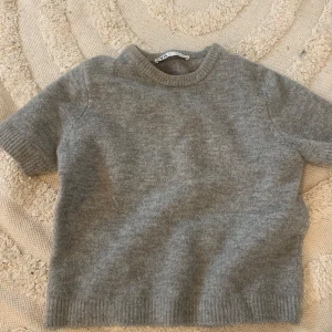 Grå stickad tröja - Mysig grå stickad tröja med korta ärmar. Perfekt för kyligare dagar och enkel att matcha med olika outfits. Tröjan har en rund halsringning och ribbade kanter för en klassisk look.