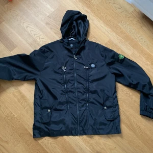 Svart vindjacka från Stone Island - Säljer en svart vindjacka från Stone Island med huva och dragkedja. Jackan har flera praktiska fickor och en ikonisk logga på ärmen. Perfekt för blåsiga dagar! Är öppen att diskutera pris!