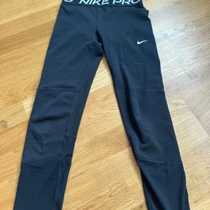 Svarta leggings från Nike - Säljer ett par svarta Nike Pro leggings med låg midja och en liten vit Nike-logga på benet. Perfekta för träning med en tight passform och elastiskt material som ger bra stöd. Midjebandet har Nike Pro-tryck för extra stil.