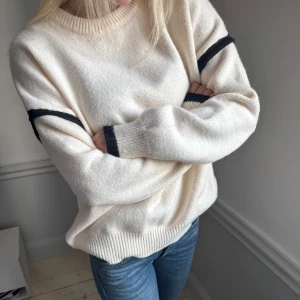Vit stickad tröja med svarta detaljer - Säljer en stilren vit stickad tröja med svarta ränder på ärmarna. Tröjan har en rund halsringning och långa ärmar. Perfekt för en avslappnad look. Hör av dig vid intresse!