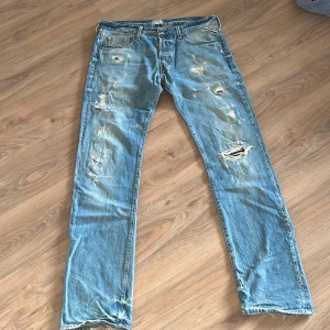 Blå jeans från Replay - Säljer ett par blå jeans från Replay med slitna detaljer och en klassisk femficksdesign. Jeansen har en knappgylf och är i en straight passform. Perfekta för en avslappnad stil.