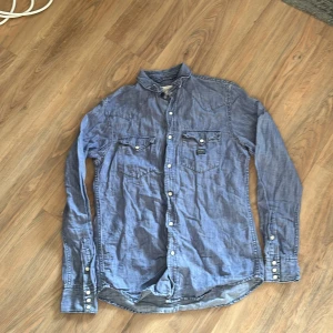 Blå jeansskjorta från Denim & Supply Ralph Lauren - Snygg blå jeansskjorta från Denim & Supply Ralph Lauren med knappar och två bröstfickor. Perfekt för en avslappnad stil. Skjortan har långa ärmar och en klassisk krage.