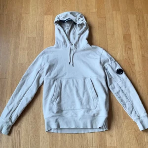 Beige hoodie från C.P. Company - Säljer en stilren beige hoodie från C.P. Company med en praktisk ficka framtill och en ikonisk linsdetalj på ärmen. Perfekt för en avslappnad look. Passar bra till både jeans och joggers.