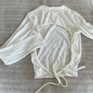 Vit ribbad topp med öppen rygg - Säljer en stilren vit ribbad topp med långärmad design och en snygg öppen rygg. Toppen har knytband i midjan för en justerbar passform. Perfekt för en trendig look!