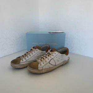 Vit begiea sneakers från Philippe Model - Snygga vita sneakers från Philippe Model med bruna detaljer. Skorna har en klassisk design med snörning. Är i väldigt gött skick bara att undersulan är lite sliten där bak på den högra skon! Pris kan diskuteras😁