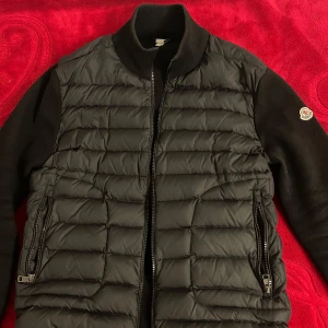 Svart moncler cardigan - Säljer en svart cardigan  från Moncler med quiltad front och stickade ärmar. Jackan har dragkedja framtill, två fickor med dragkedja och ribbad krage. Klassisk Moncler-logga på ärmen. Perfekt för dig som vill ha en stilren och sportig look.