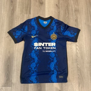 Inter Milan blå matchtröja Nike - Inter Milan fotbollströja i blått med svart och ljusblått ormmönster, officiell klubbmärke och sponsortryck på bröstet. Kortärmad modell med svart krage och ärmslut. Tillverkad i lätt och ventilerande material, perfekt för match eller träning. Storlek S (passar också M)