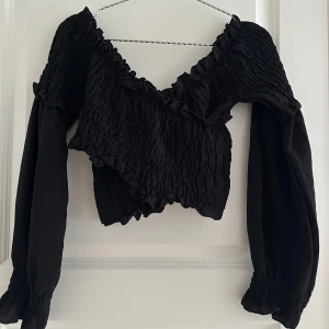 Svart croppad blus med volang - Supersnygg svart croppad blus från Bershka med långärmade puffärmar och smockad struktur. Toppen har v-ringning och volangdetaljer längs kanterna för en trendig look. Perfekt att styla med höga jeans eller kjol.