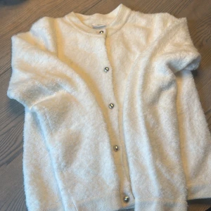 Vit fluffig kofta med silver knappar - Supermjuk och fluffig vit kofta från Kappahlmed silver knappar framtill. Långärmad och rak passform, perfekt att slänga på sig när det är kyligt. Koftan har en enkel, stilren look och är gjord i ett stickat material som känns riktigt cozy.