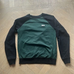 Grön och svart sweatshirt från Vans - Snygg crewneck från Vans i mörkgrönt med svarta ärmar och detaljer. Tröjan har rund halsringning, ribbade muddar och en liten Vans-patch på bröstet. Perfekt för dig som gillar streetwear och vill ha en stilren look.