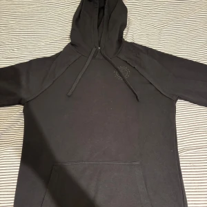 Svart hoodie från Gymshark M - Svart hoodie från Gymshark i storlek M. Klassisk modell med huva och dragsnören, samt en stor magficka framtill. Tillverkad i mjuk bomullsmix, perfekt för chill eller gym. Diskret Gymshark-logga på bröstet och ribbade muddar vid ärmslut. Har använts fåtal gånger under träning.