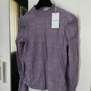 Lila smockad blus från Vila - Supersöt lila blus från Vila med smockad struktur och hög hals. Långa ärmar med lätt puff vid axlarna och smockade muddar. Perfekt för dig som gillar en trendig och romantisk look. Färgen är en mjuk pastellila som verkligen poppar. Helt ny, men var inte riktigt min stil därav säljer jag den. Nypris är 399kr men jag säljer den för endast 250kr💓