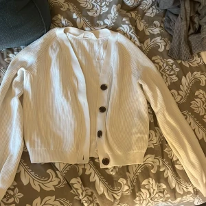 Vit ribbad kofta från H&M - Säljer en vit ribbad kofta från H&M med stora svarta knappar framtill. Koftan har lång ärm, normal passform. Strl 158/164