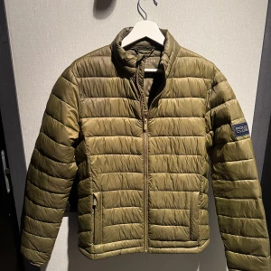 Olivgrön pufferjacka från Polo Club - Snygg olivgrön pufferjacka från Polo Club med quiltad design och dragkedja framtill. Jackan har ståkrage, två fickor och en badge på ärmen. (OBS, lite missfärgad åå höger axel, se bild 3.) 