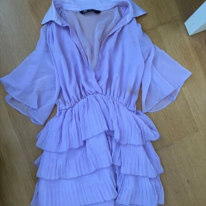 Ljuslila volangklänning från Zara - Superfin ljuslila klänning från Zara med omlottliv, v-ringning och flera lager volanger i kjolen. Klänningen har vida, korta ärmar och är gjord i ett lätt, flowy material!