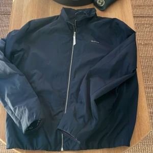 Mörkblå vindjacka från GANT - Snygg mörkblå vindjacka från GANT med hel dragkedja framtill och diskret logga på bröstet. Jackan är tillverkad i 100% polyester och har en stilren, sportig look med hög krage och långa ärmar. Perfekt för dig som gillar klassisk och clean stil.