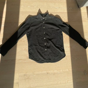 Svart från L'Homme Rouge - Svart skjorta från L'Homme Rouge med klassisk krage, vita knappar och bröstficka. Skjortan har lång ärm och en lite skrynklig textur som ger en avslappnad vibe. Perfekt för dig som gillar stilrena och enkla plagg. Mottagaren står för frakten. Priset är sänkt från 299 till 199kr