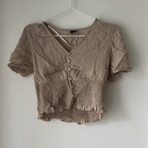 Beige blus från Vero Moda - Supersöt beige blus från Vero Moda med korta ärmar, v-ringning och knappar framtill. Nedre delen är smockad med volangkant för en extra chill vibe. Perfekt till jeans eller kjol när du vill ha en avslappnad men ändå snygg look.