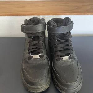 Svarta Nike Air Force 1 Mid sneakers - Klassiska helsvarta Nike Air Force 1 Mid med snörning och kardborreband runt ankeln. Skorna är i skinn med rund tå och platt sula. Perfekta för dig som gillar en clean och tidlös streetstil. Swoosh-logga på sidan och Nike Air-detaljer på hälen.
