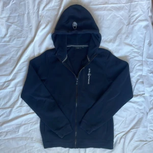 Svart hoodie från Sail Racing - Snygg svart hoodie från Sail Racing med dragkedja och broderad logga på bröstet. Tröjan har huva, lång ärm och en praktisk bröstficka med dragkedja. Mjuk insida och stilrena vita detaljer vid huvkanten. Perfekt för kyliga dagar.